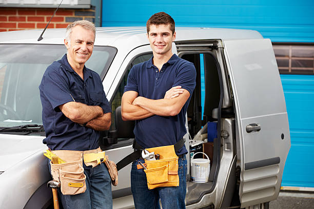 Two men in matching blue shirts and tool belts standing confidently beside a van with its door open, ready for work.